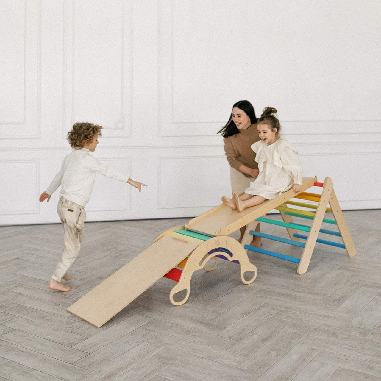 Children playing on a Montessori climbing set with rainbow-colored rungs and slide indoors.