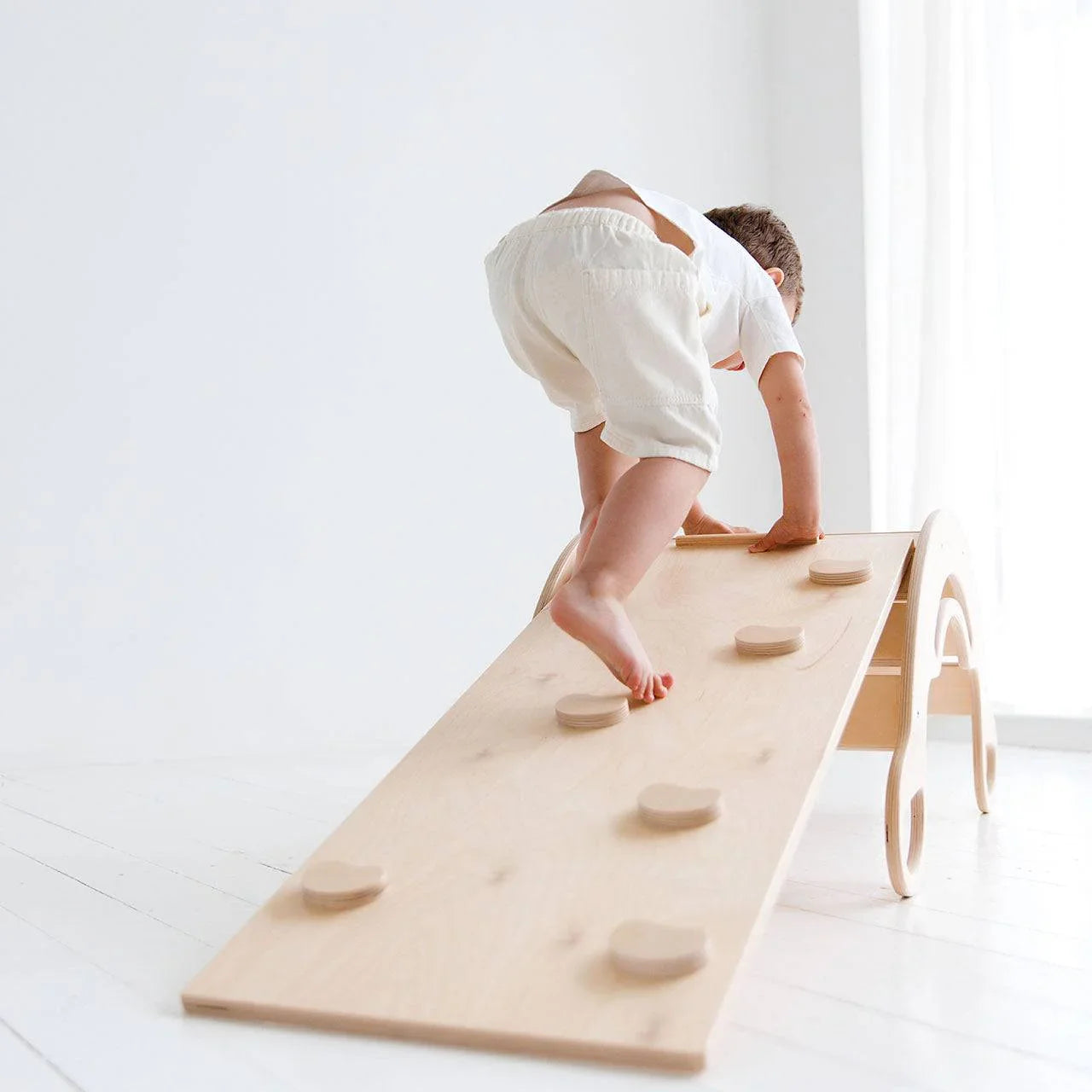 Child climbing on a natural wood Montessori rocker with dual-sided board promoting motor skills development.