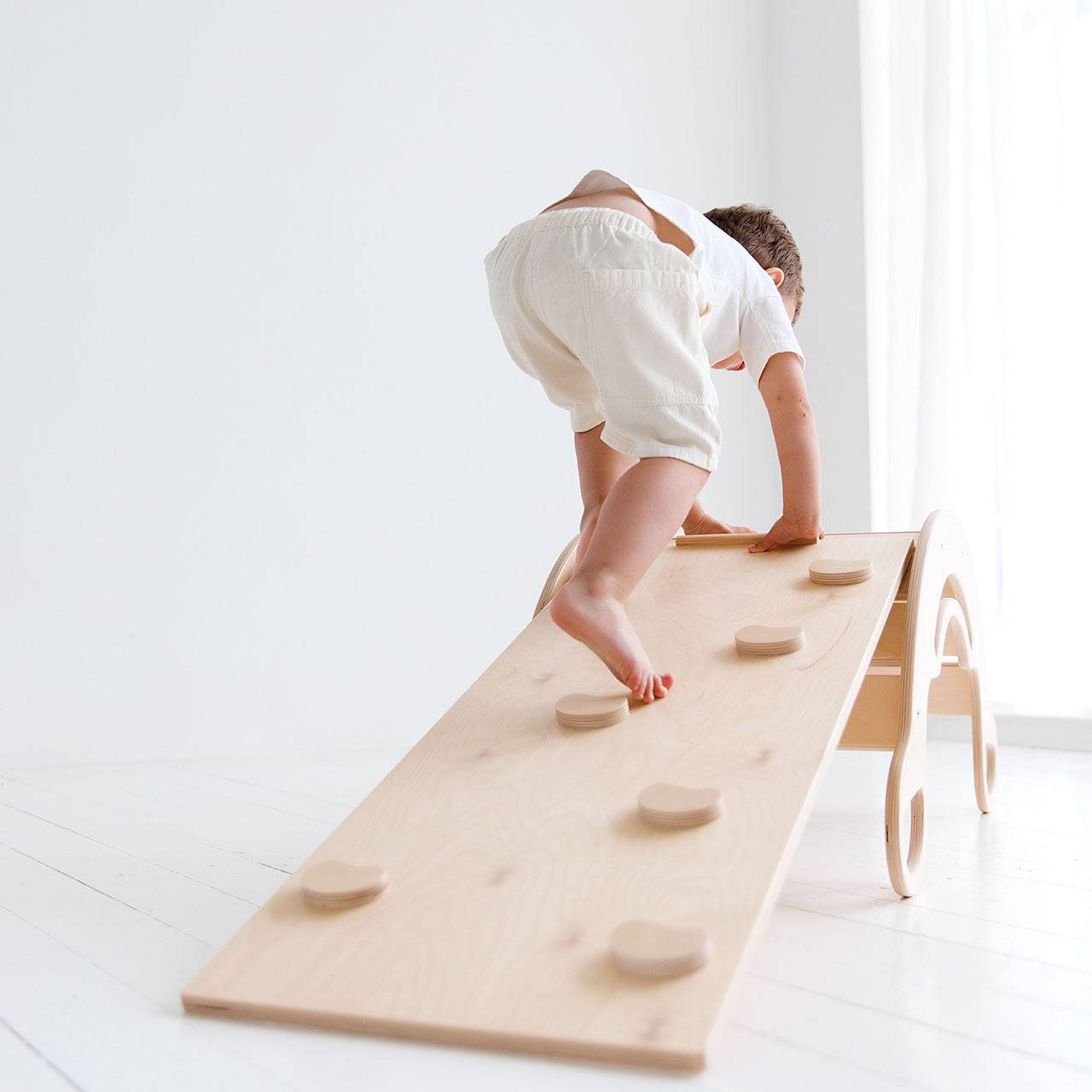 Child climbing on a natural wood Montessori rocker with dual-sided board promoting motor skills development.