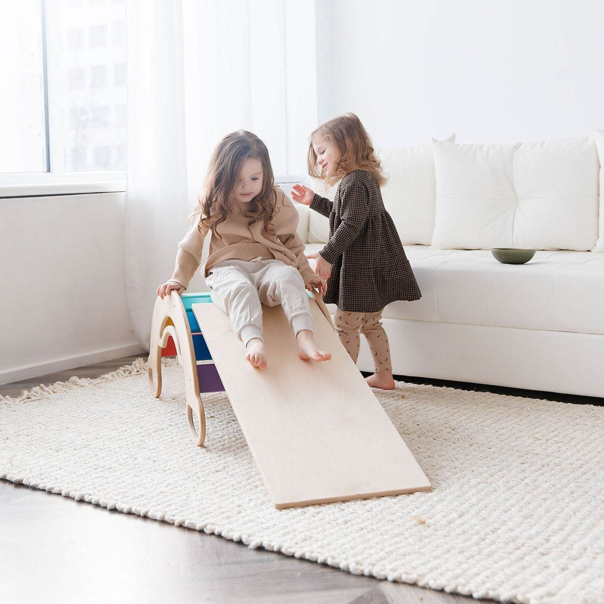 Children playing on a wooden rocker and double-sided board, part of a light-colored set with a mini chair, in a cozy living room.