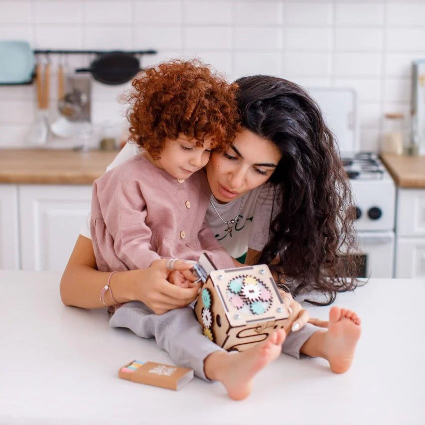 Toddler and adult playing with interactive wooden activity cube in kitchen.