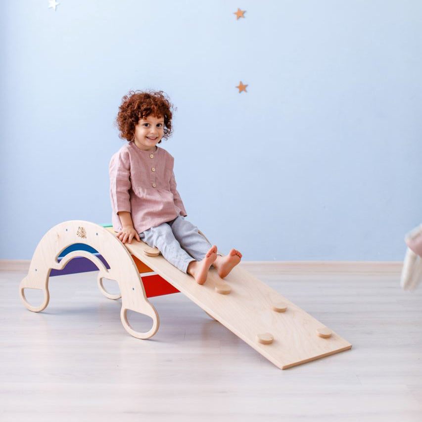 Child playing on a wooden seesaw set with slide and mini chair, colorful background, eco-friendly design.
