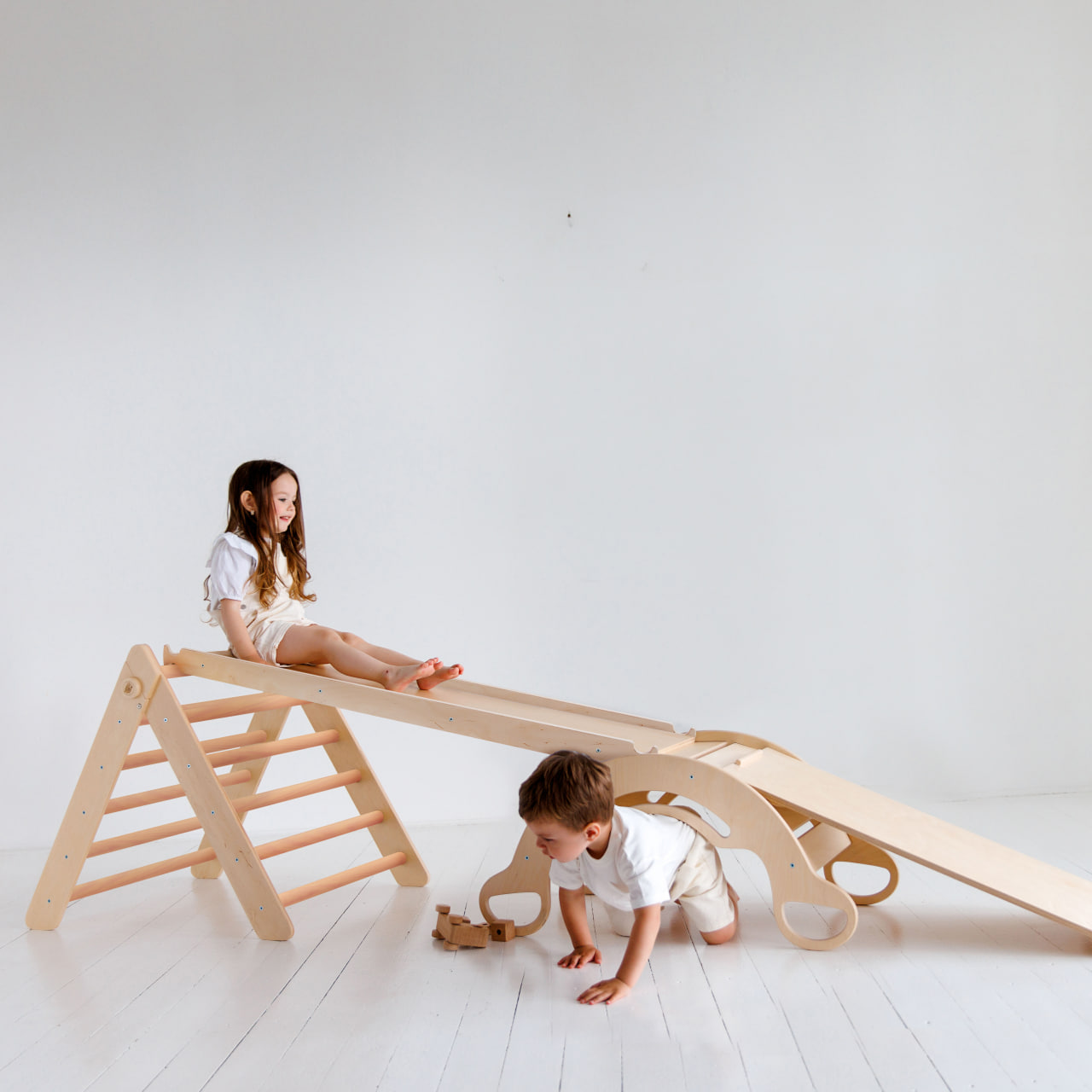 Children playing on a natural wooden Pikler triangle set with slides and a balance rocker.