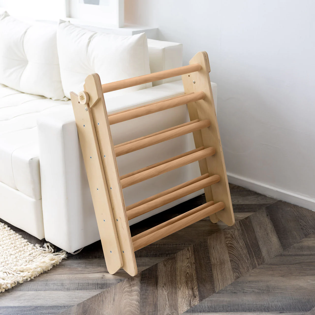 Pikler triangle climbing set with a wooden ladder placed next to a white sofa on a wooden floor.