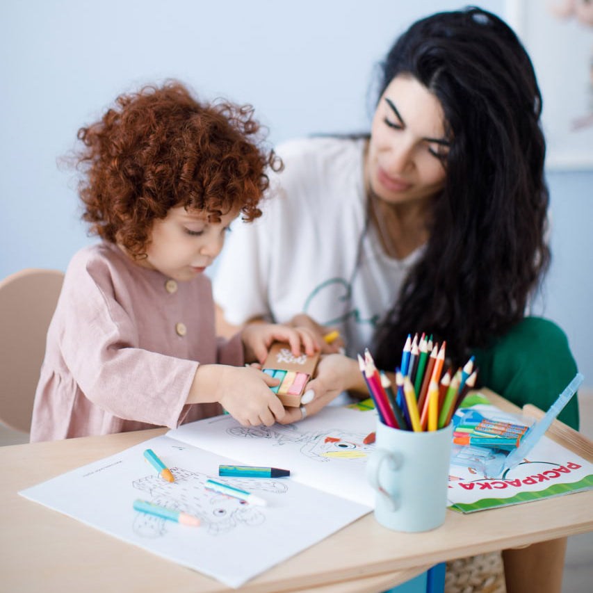 Child using toy blocks on a table with adult assistance, featuring coloring pages and pencils.