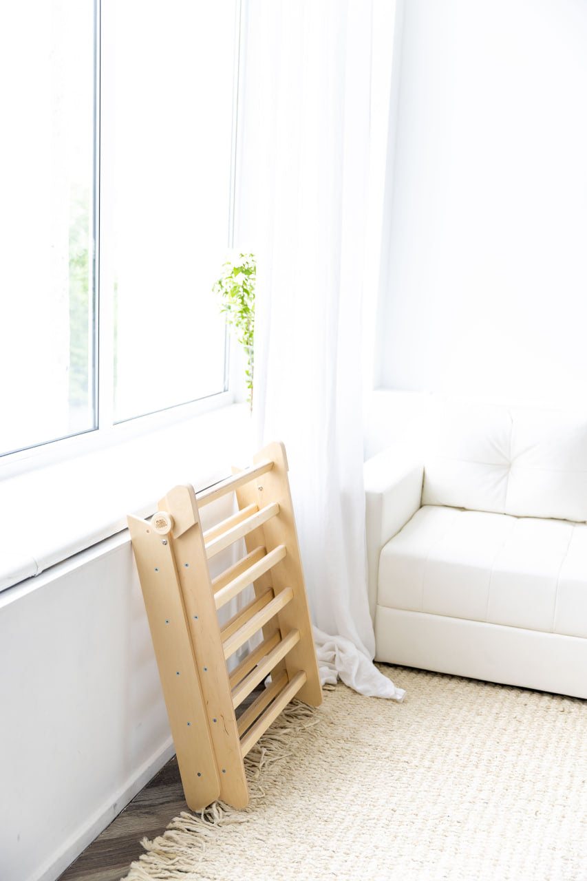 Natural wood climbing triangle set with slide/ladder for kids, folded beside a window.