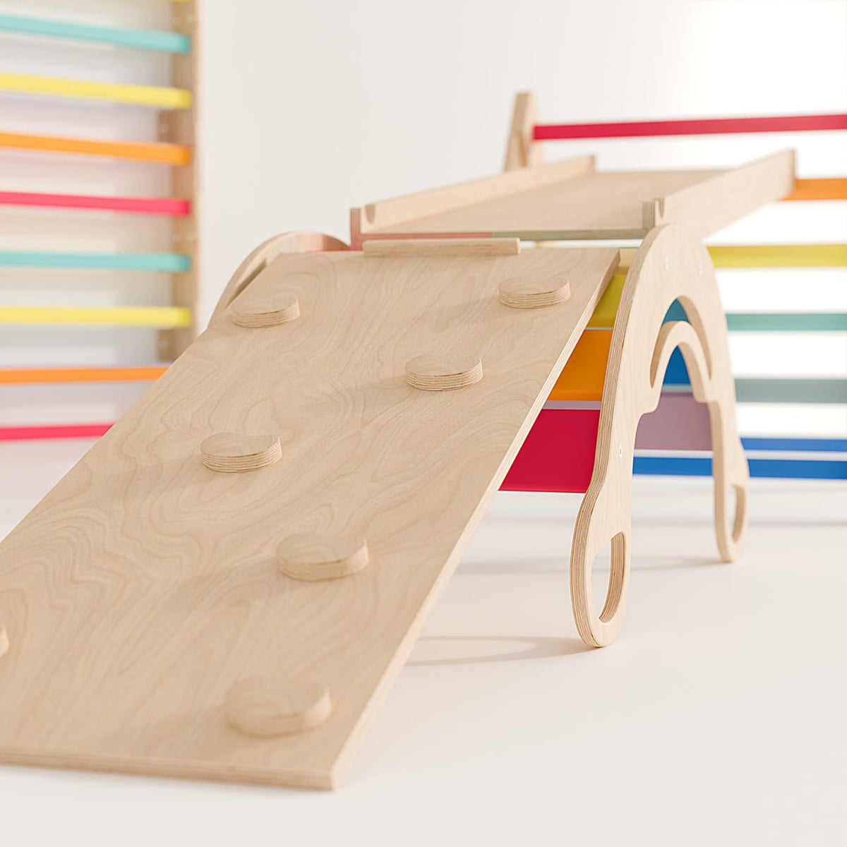 Indoor children&#39;s play set featuring a Sprossenwand, Pikler triangle, slides, and balance rocker in rainbow colors.