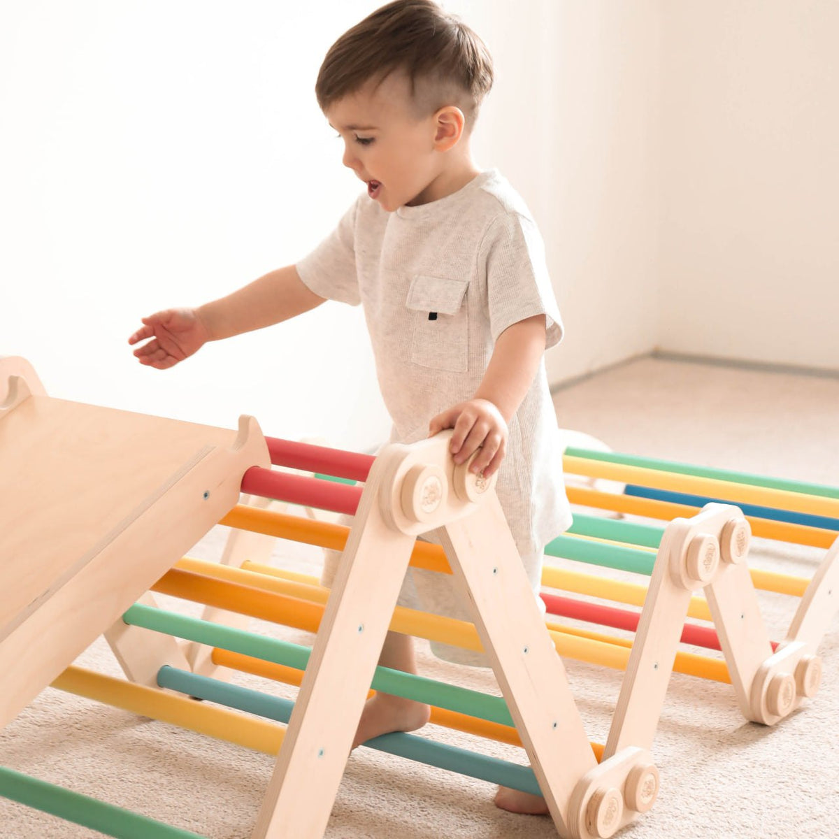 Child playing on rainbow-colored climbing frame with slide and Pikler triangles, promoting fun and motor skills.