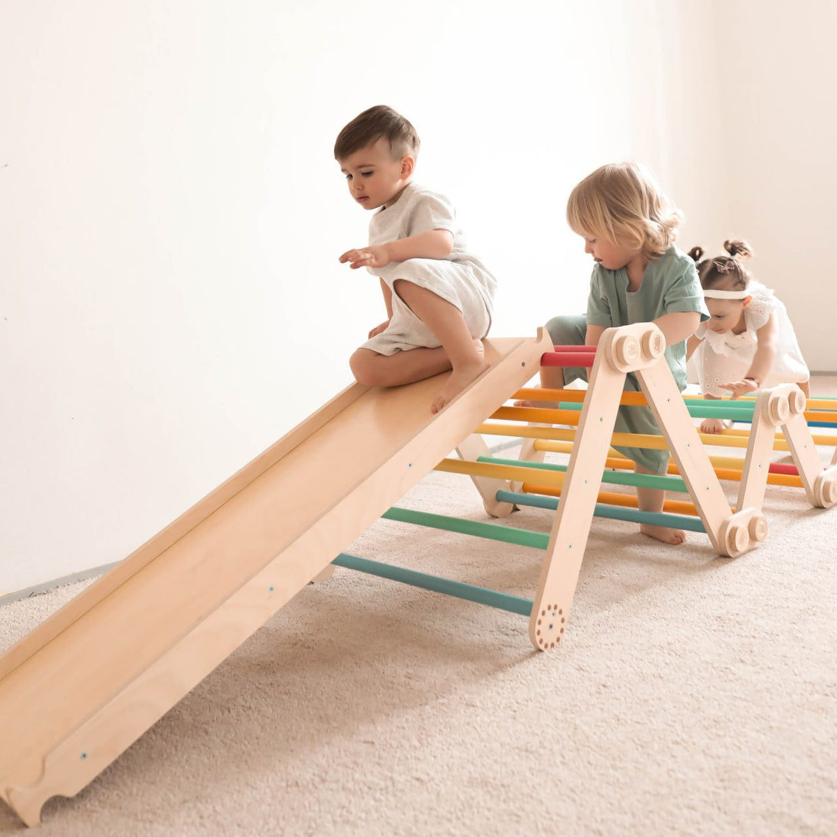 Children playing on a rainbow-colored wooden climbing frame set with a slide indoors.