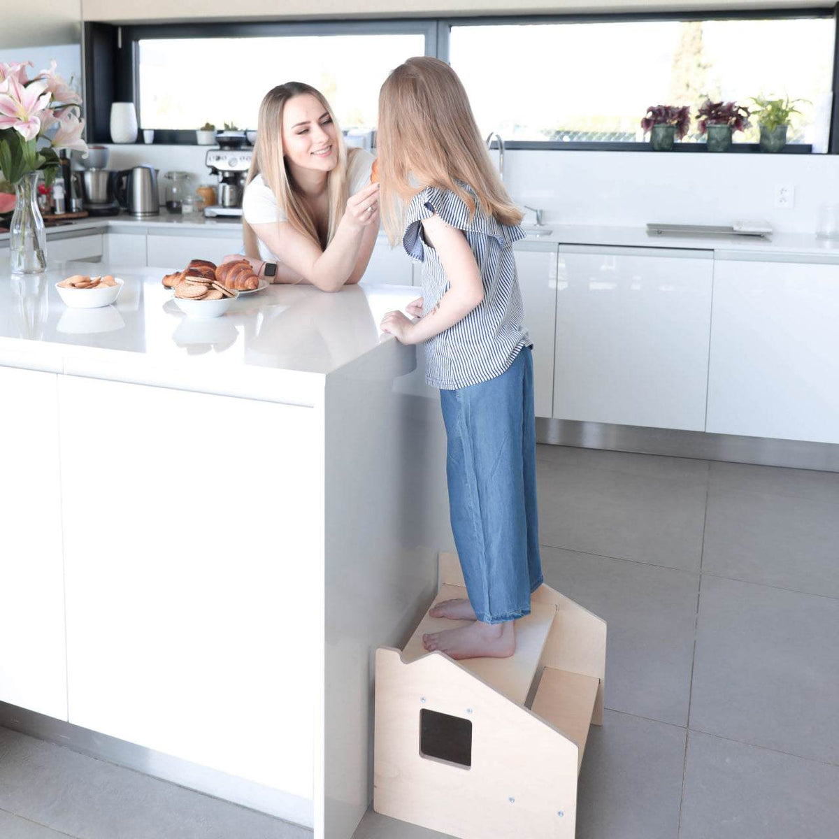 Child using Montessori step stool in modern kitchen to reach counter, emphasizes safety and independence.