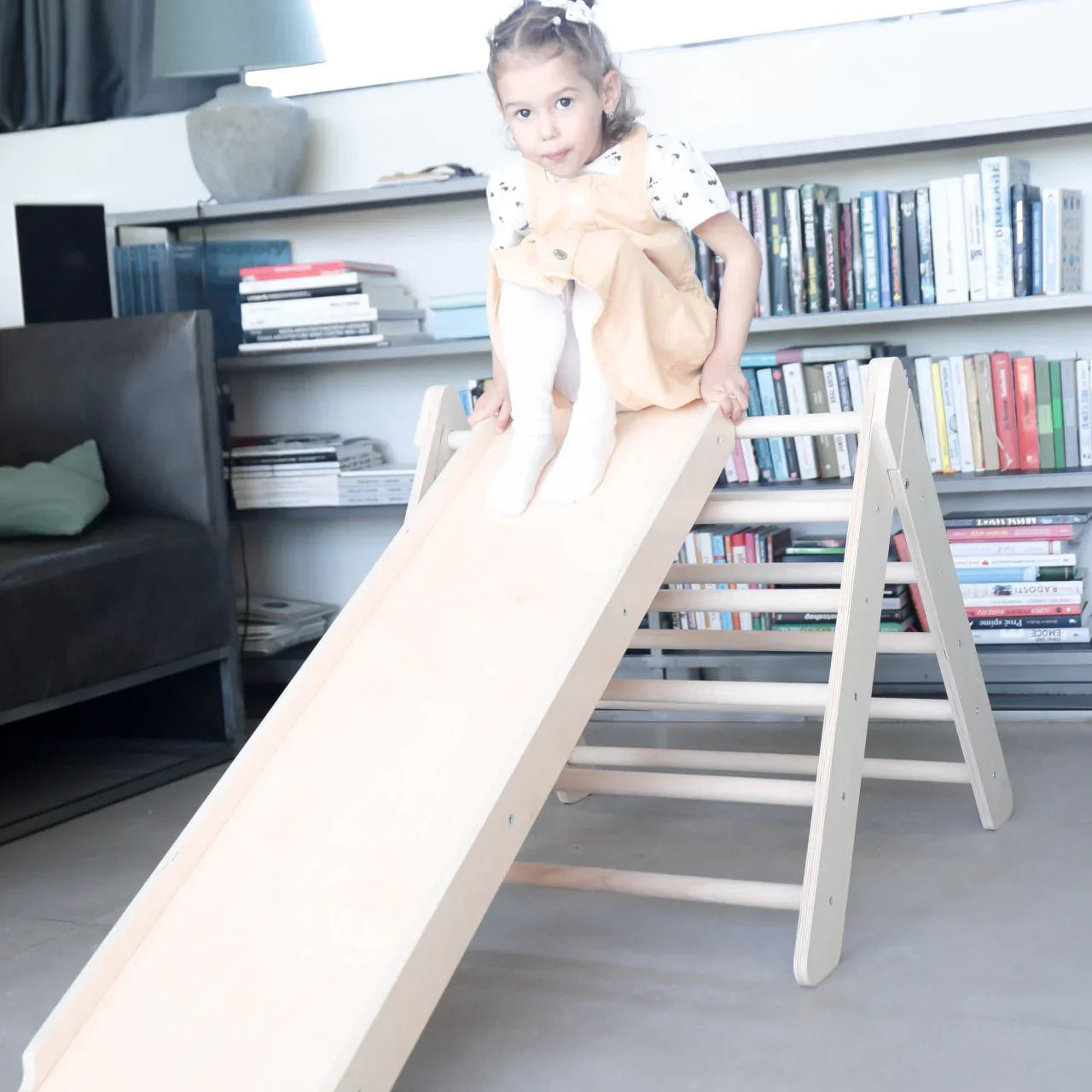 Child playing on an uncoated wooden Pikler triangle and slide in a living room setting.