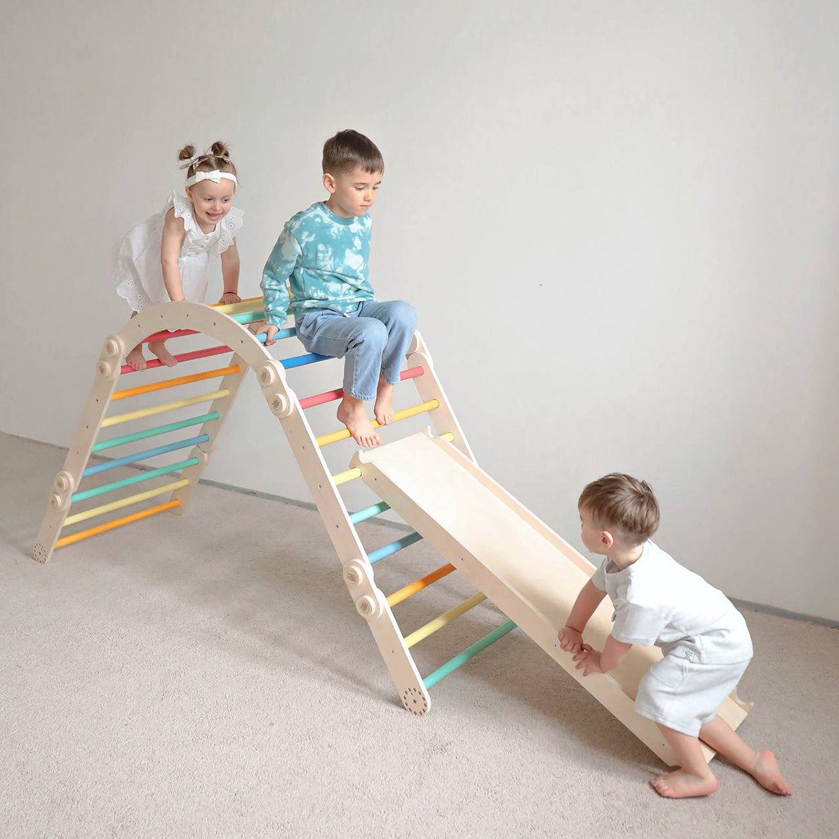 Kids playing on rainbow-colored wooden climbing set with slide indoors.
