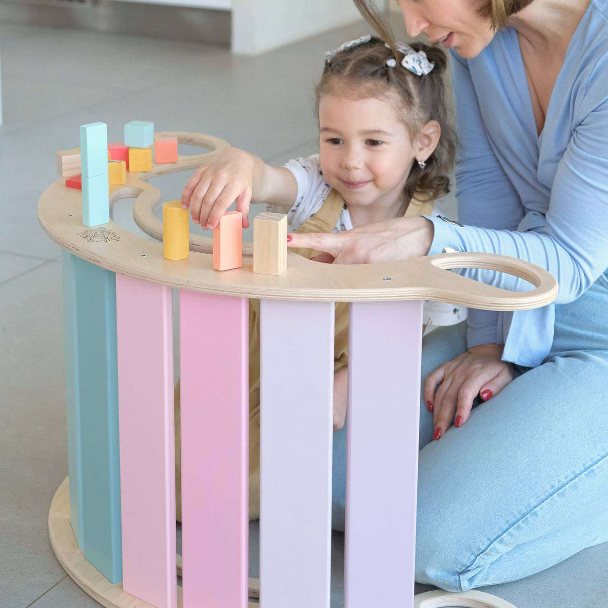 Balancewippe in Pastellfarben mit spielendem Kind und Erwachsener, vielseitig als Schaukel, Sitzgelegenheit oder Klettergerüst verwendbar.